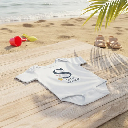Baby onesie with 'S is for Samuel' text on a wooden surface by the beach.