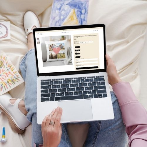 Woman sitting comfortably and customizing a personalized photo pillow on an online store using her laptop, surrounded by artwork and stationery.